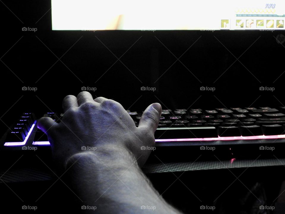 Teenager boy playing Minecraft on the gaming computer seen from his left side and close-up of his hand playing in a dark room.