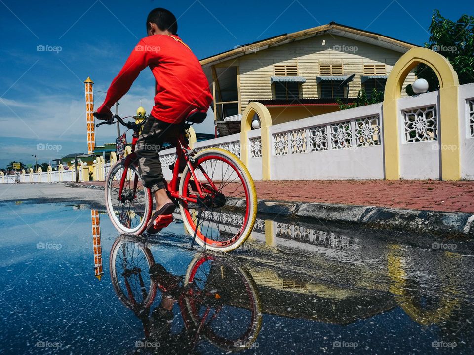 A boy riding the bicycle in a puddle infront of a mosque under bright sunny day