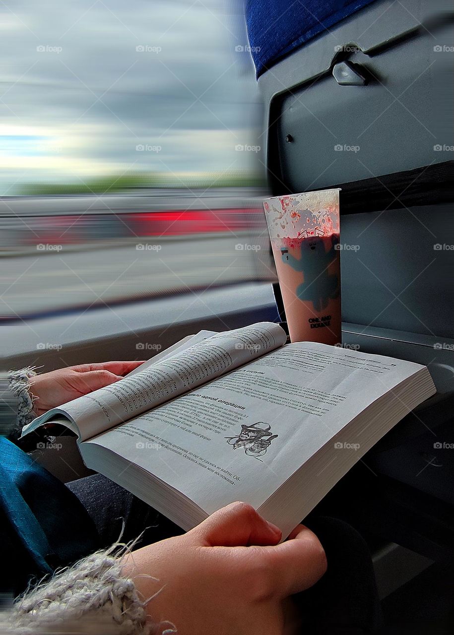 In the train car. Hands hold a book, which lies on a folding stand. Nearby is a transparent glass of coffee. Blurred landscape outside the train window