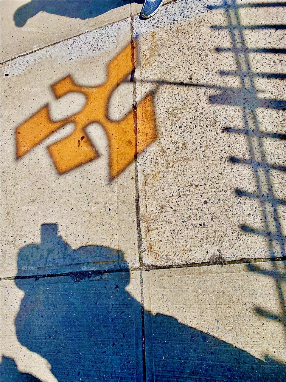An orange shape like a piece to a jigsaw puzzle is cast on the sidewalk along with the shadow of a fence, the photographer, a metal pole and an attachment for the shape so it can spin when blown around by the wind. 2022. Hypnotic Productions