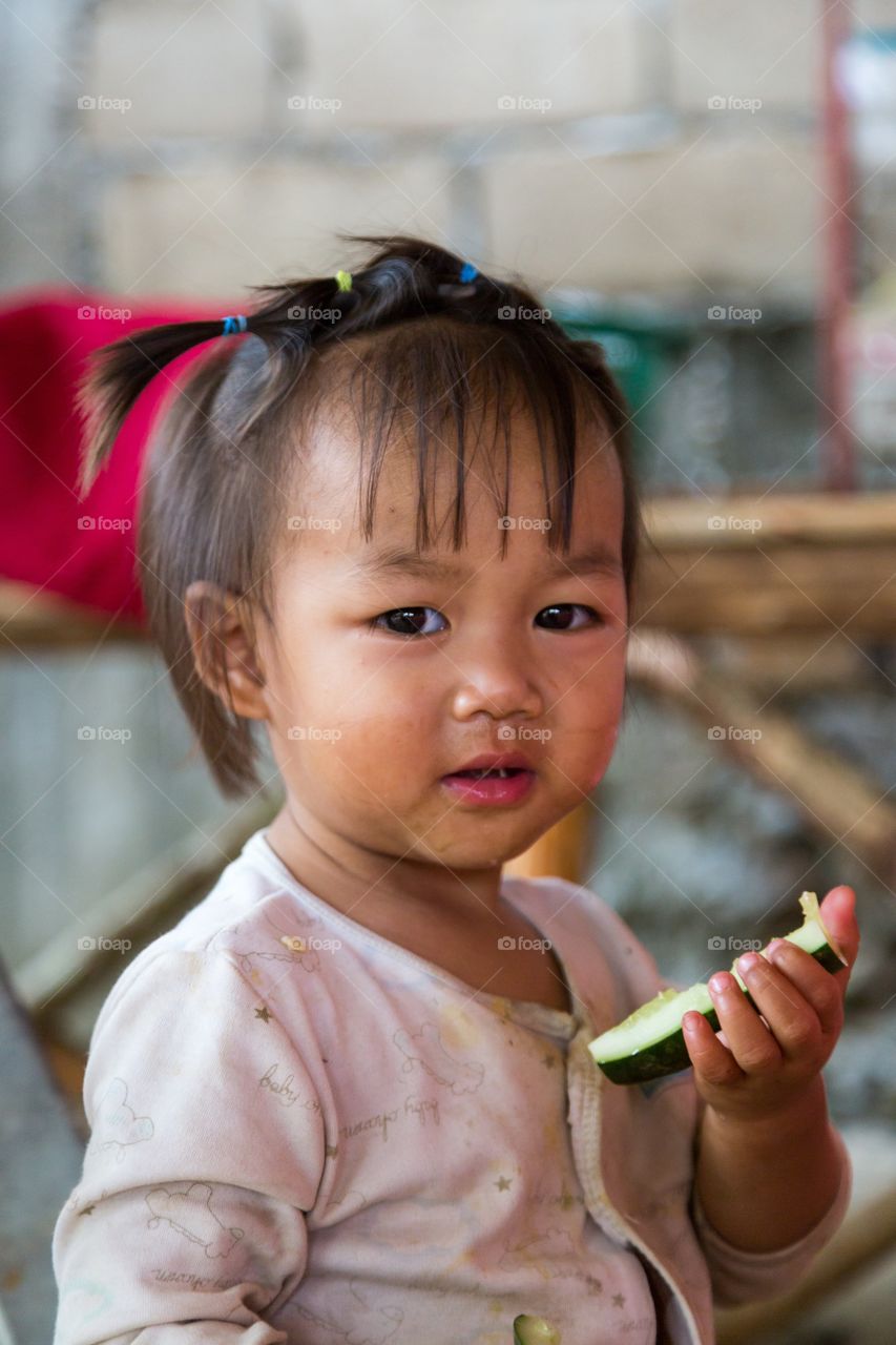 Little girl eats cucumber. Small girl from Laos eats cucumber. Out of focus background. Shy smile. Cute. Ponytails