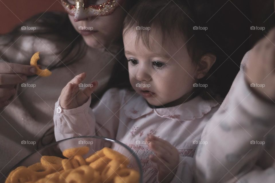 Portrait of a beautiful caucasian baby girl with sisters eating corn rings with a transparent glass dish and enthusiastically with real emotions watching a cartoon on a tablet while sitting together on a sofa, close-up side view. Family watching movi
