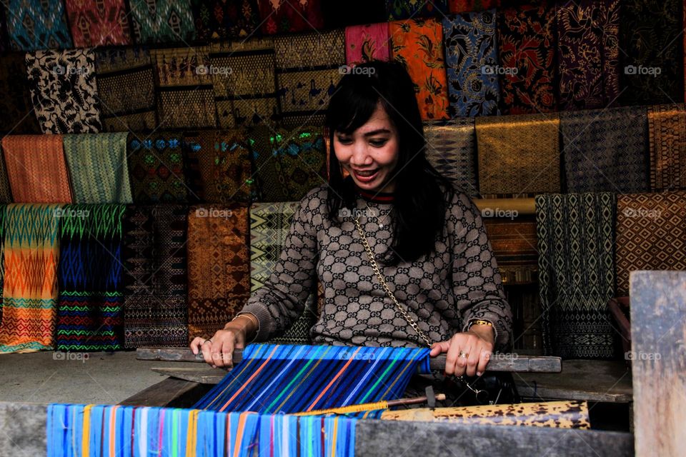 Portrait of a young woman sitting learning to weave cloth with traditional tools at a traditional cloth craft workshop in Sade village, with a happy expression, Lombok, Indonesia.