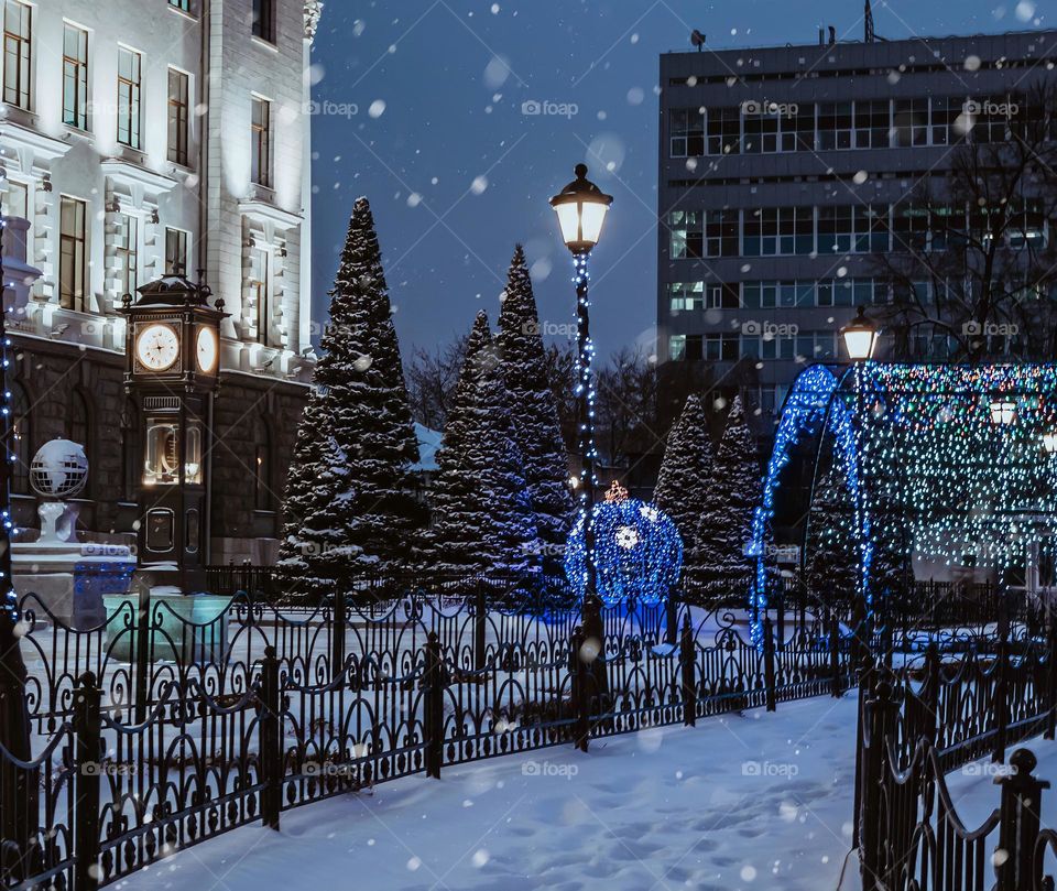 Beautiful city night winter landscape. Tower clock on the square and snow-covered firs
