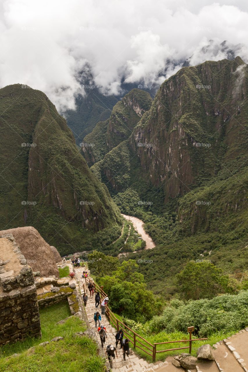View towards deep valley. View from Machu Picchu towards deep valley. Steep mountains. River at the bottom. People climbing up. Cloudy sky
