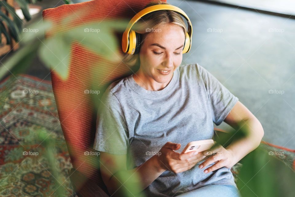 Young smiling blonde woman with close eyes in yellow headphones enjoys music with mobile phone sitting on chair at cafe