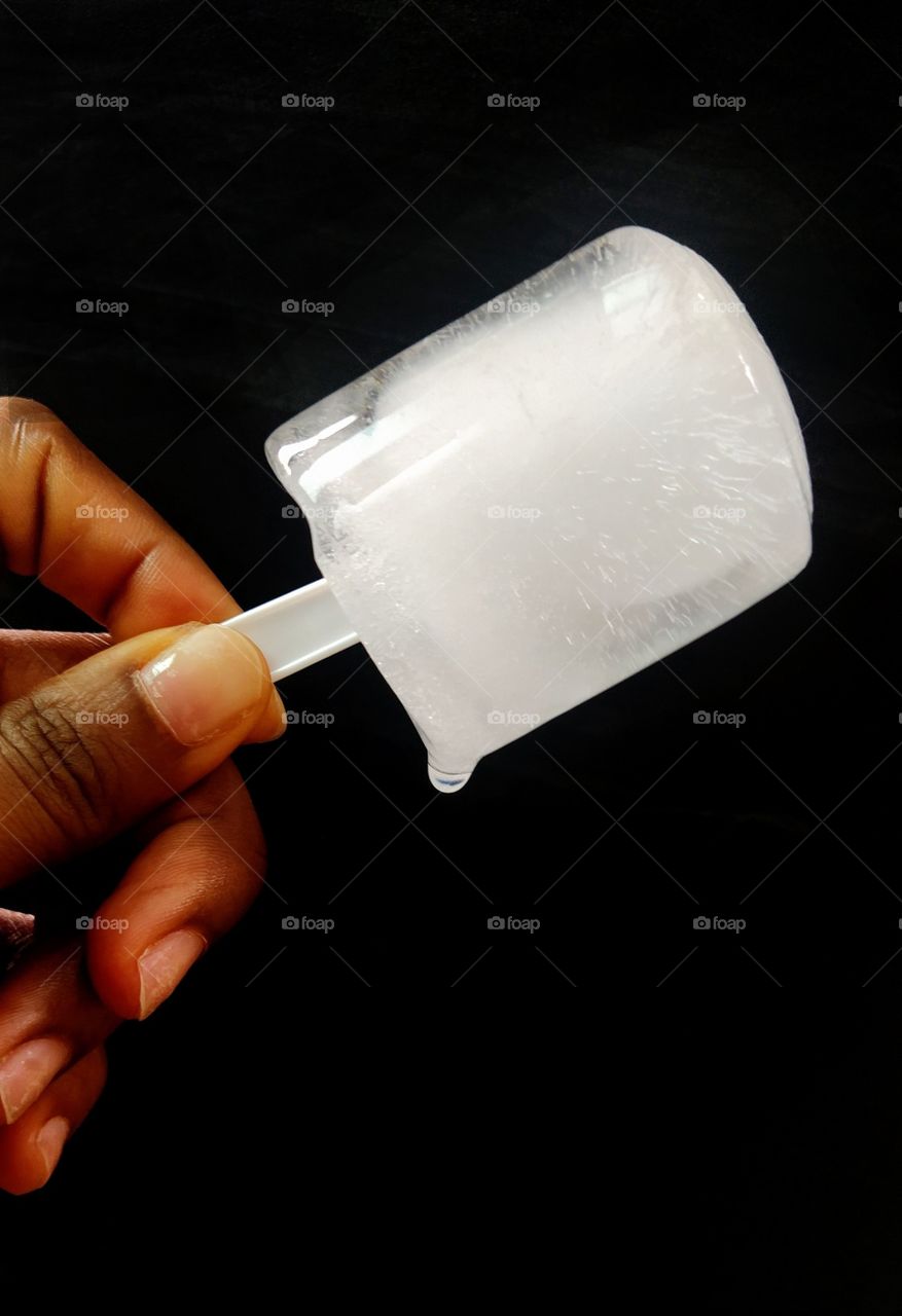 A hand holding a dripping cylindrical shaped ice block on a stick