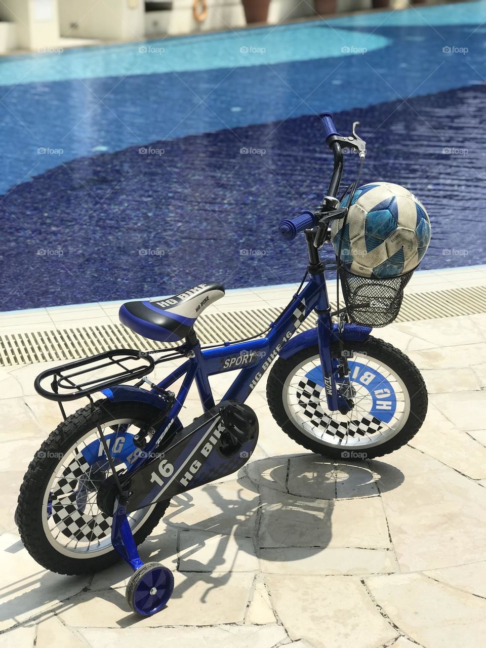 A blue bicycle has a blue football in it’s front basket and stands near blue water swimming pool 😝