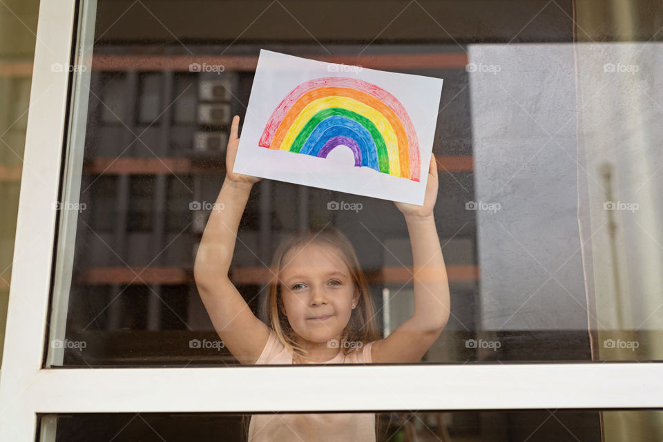 Happy little Caucasian girl with blonde hair painting rainbow during COVID-19 coronavirus quarantine and self isolation 