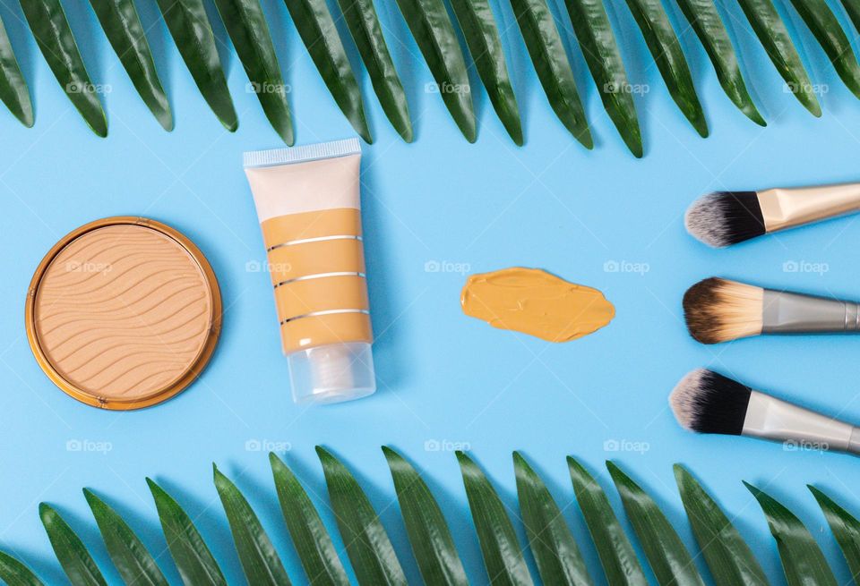 A tube of foundation,a round face powder,three makeup brushes and a smear of foundation lie in a row on a blue background with a palm tree branch around the edge,flat lay. Concept female cosmetics, beauty salon.