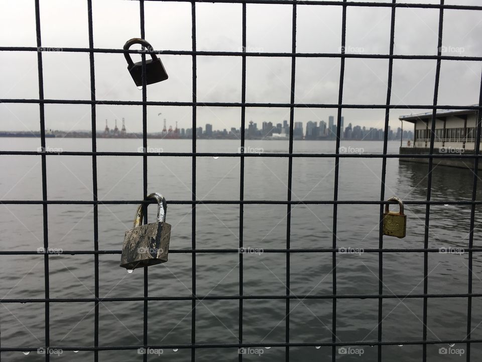 Locks on a fences near a pier on a rainy day. A cityscape appears in the background. Each lock symbolizes a relationship, a love, permanent and visible. The key is thrown into the water after the lock it placed, symbolizing “forever”.