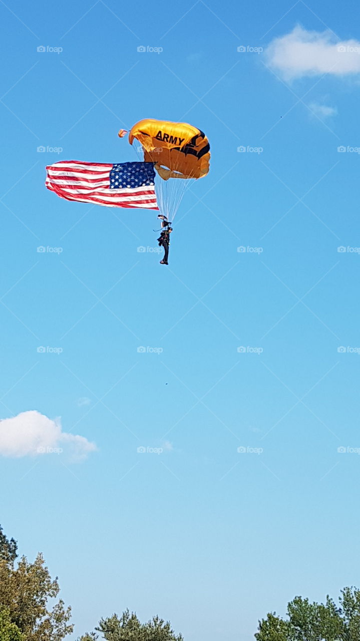 US Army Golden Knights parachuting with large American Flag in bright blue sky.