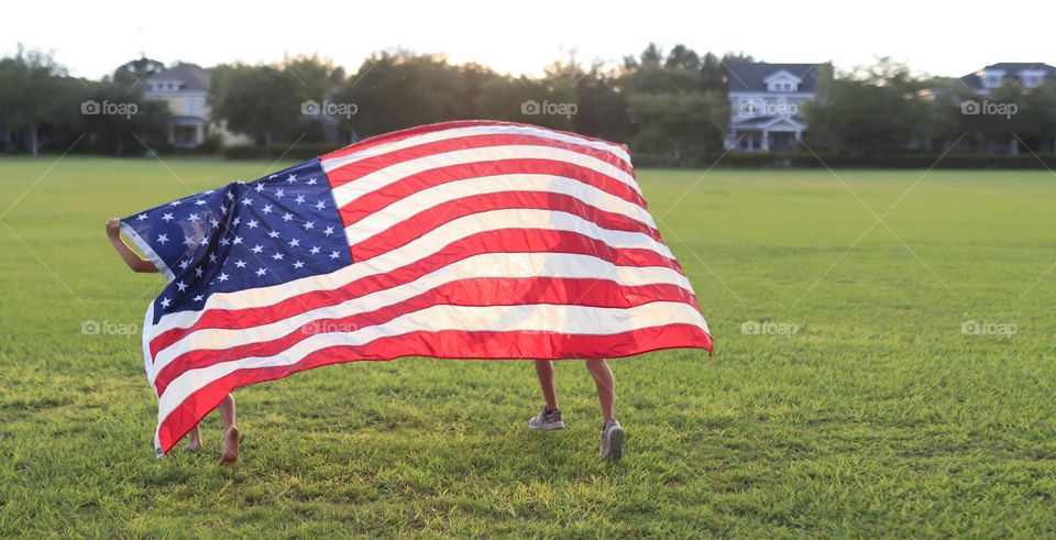 Two girls holding America flag and running on green field during sunset.