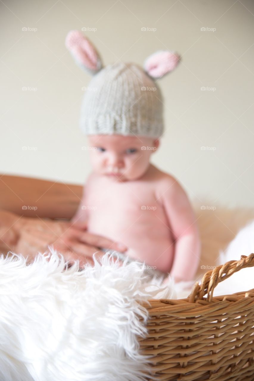 Setting up a photo shoot with a defocused newborn baby wearing bunny ears sitting in a basket with white fur - meant to illustrate the effort that goes on before the final photo is taken 