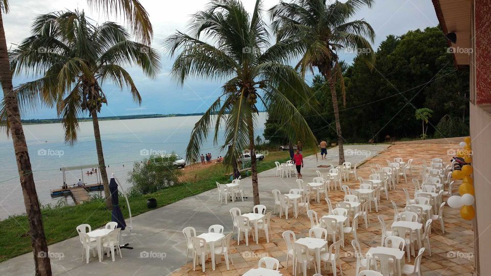 Bar, cadeiras, mesas e pessoas à beira da praia e belos coqueiros.