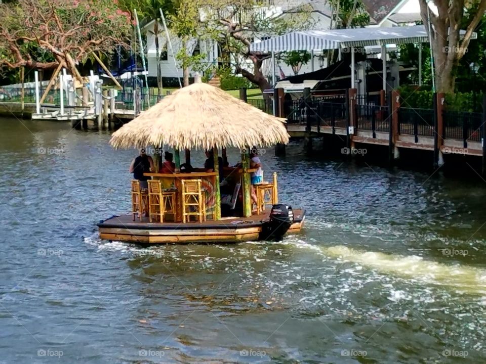 River boat round raft with engine going down river. Gated home in background
People are drinking, sitting on bar stools. Thatched roof round raft transportation system. Funny boating pic.