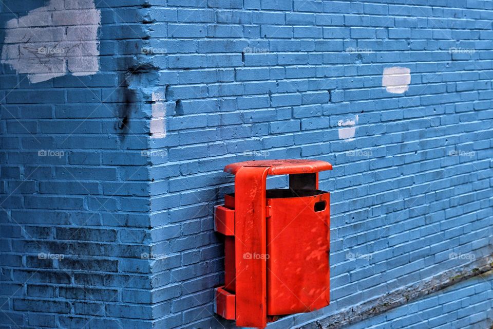 An orange trash can hangs on a blue weathered brick wall
