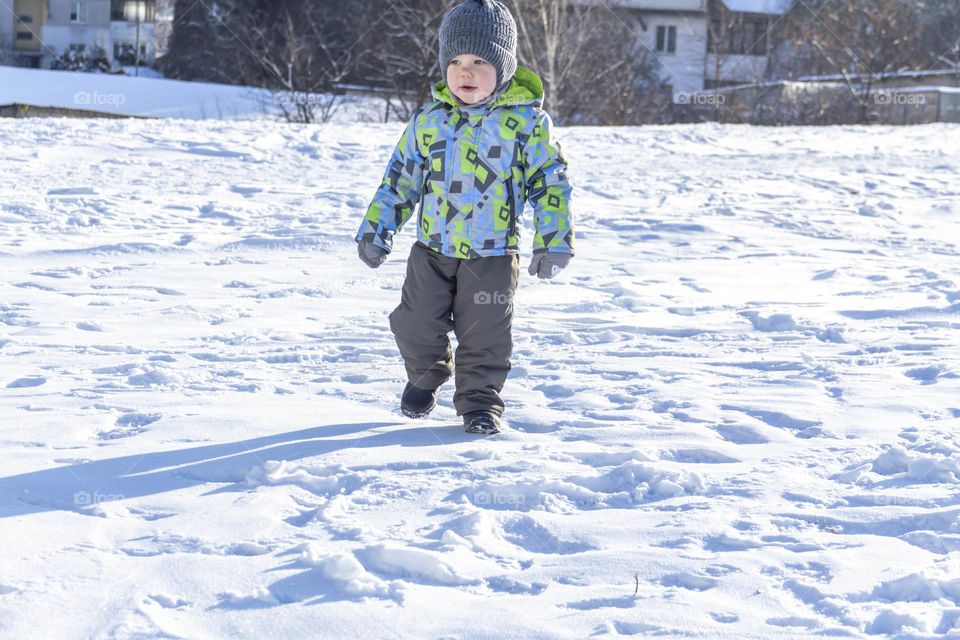 A child in winter in winter jackets, pants, a hat and boots on white snow on the street and in the park in nature plays winter fun and sleds.
