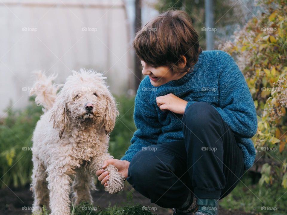 Cute little smiling boy with dark hair wearing blue knitted sweater with white curly poodle in garden, pod child 