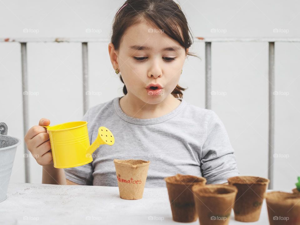 Portrait of a beautiful little caucasian brunette girl with a surprised emotion, a yellow watering can in her hands looking at craft glasses wet from watering, sitting at a stone table in the backyard of the house, close-up side view. The concept of