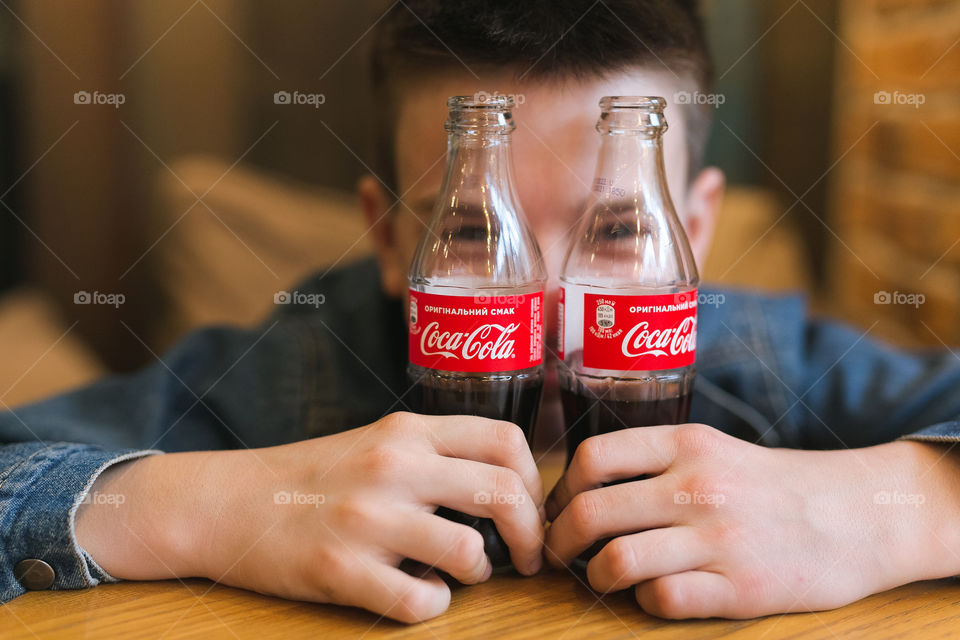 a cheerful, cute guy of fifteen years old, sitting in a cafe, smiling, having fun and looking through two bottles of Coca-Cola.  dressed in denim, charismatic teen