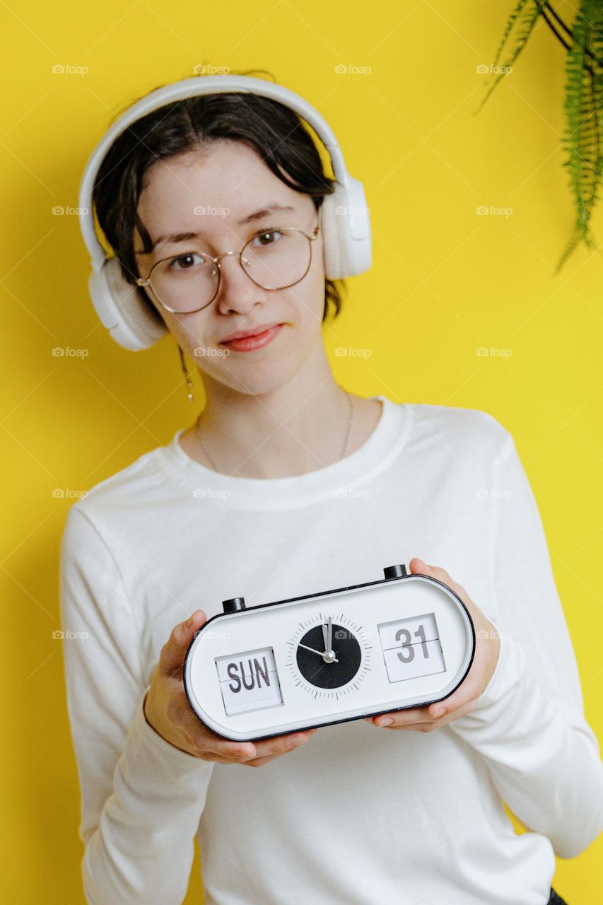 Portrait of one beautiful Caucasian happy teenage girl in white headphones with a watch in her hands on which the date and day are indicated, looking at the camera and standing against the background of a yellow wall on a summer sunny day, side view