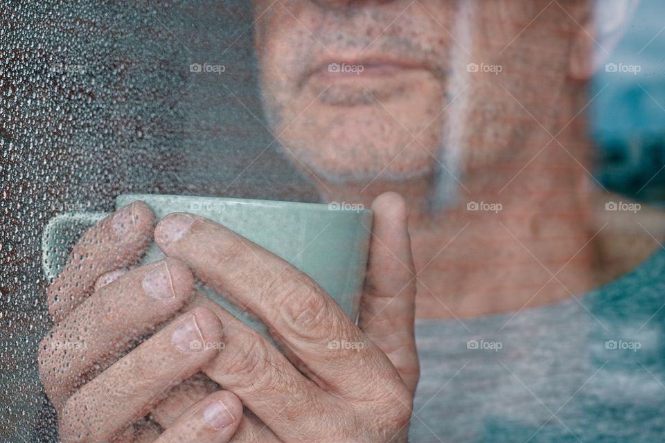 Man looking through the window on a rainy day holding a mug