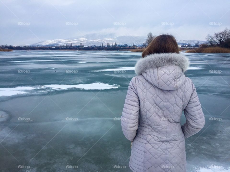 Back of woman wearing winter jacket near a frozen lake