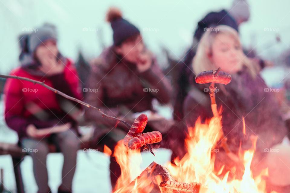 Family spending time together around campfire outdoors in the winter. People roasting sausages over the campfire using wooden sticks. People out of focus