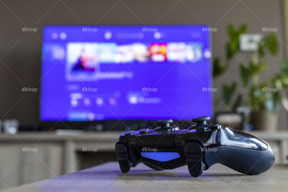 A portrait of a ps4 controller in front of a tv displaying the ps4 home screen. My spot in the house for playing a video game.
