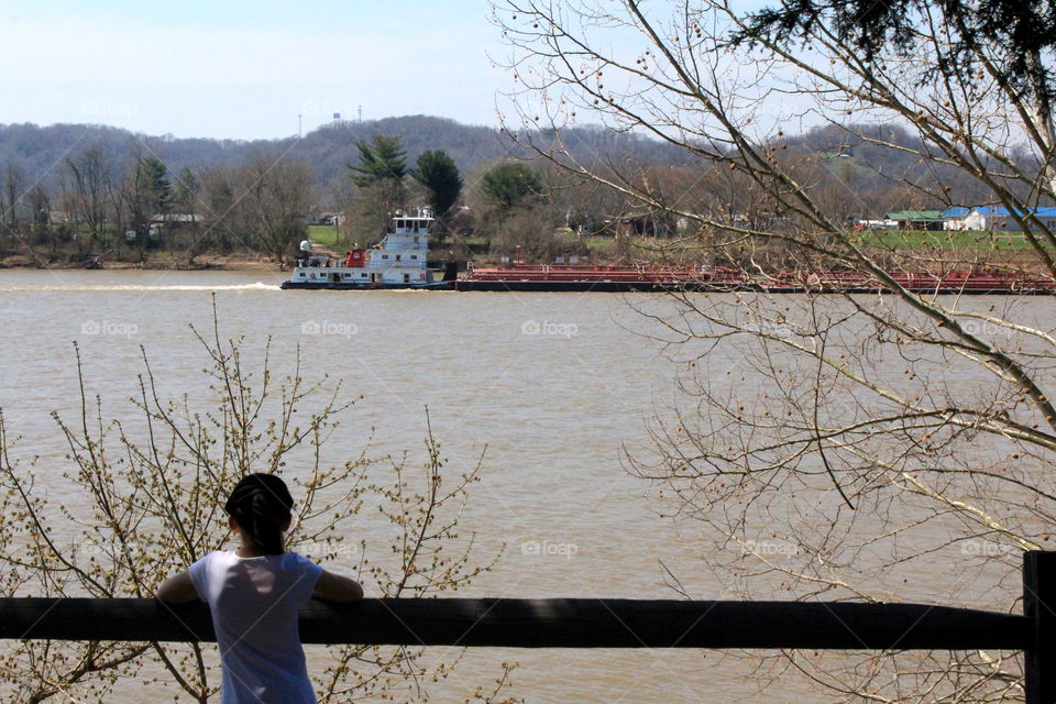 This is a picture of a little girl standing by the wood rail looking at a barge floating on the Ohio River.