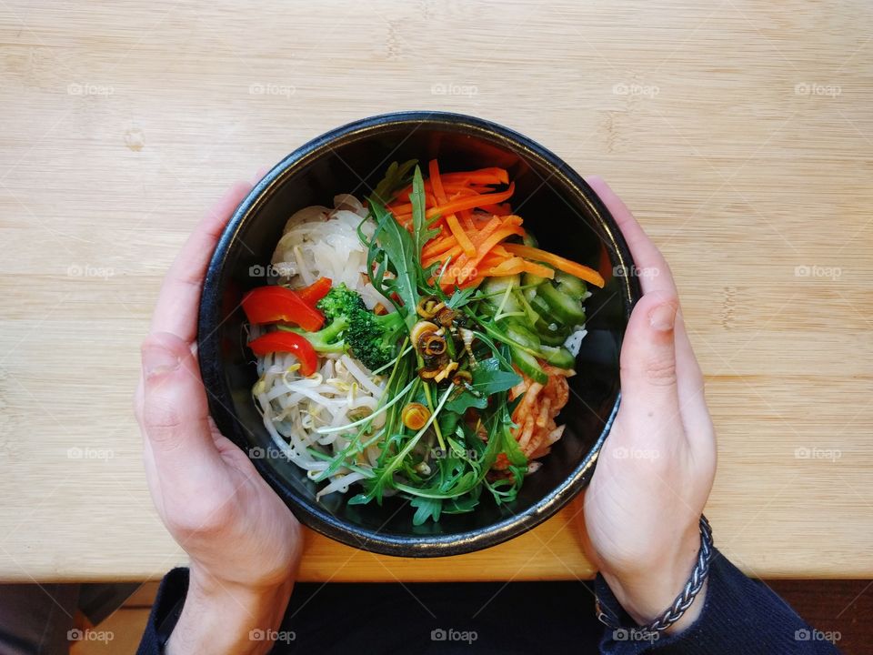 Hands holding Asian noodles and vegetables bowl