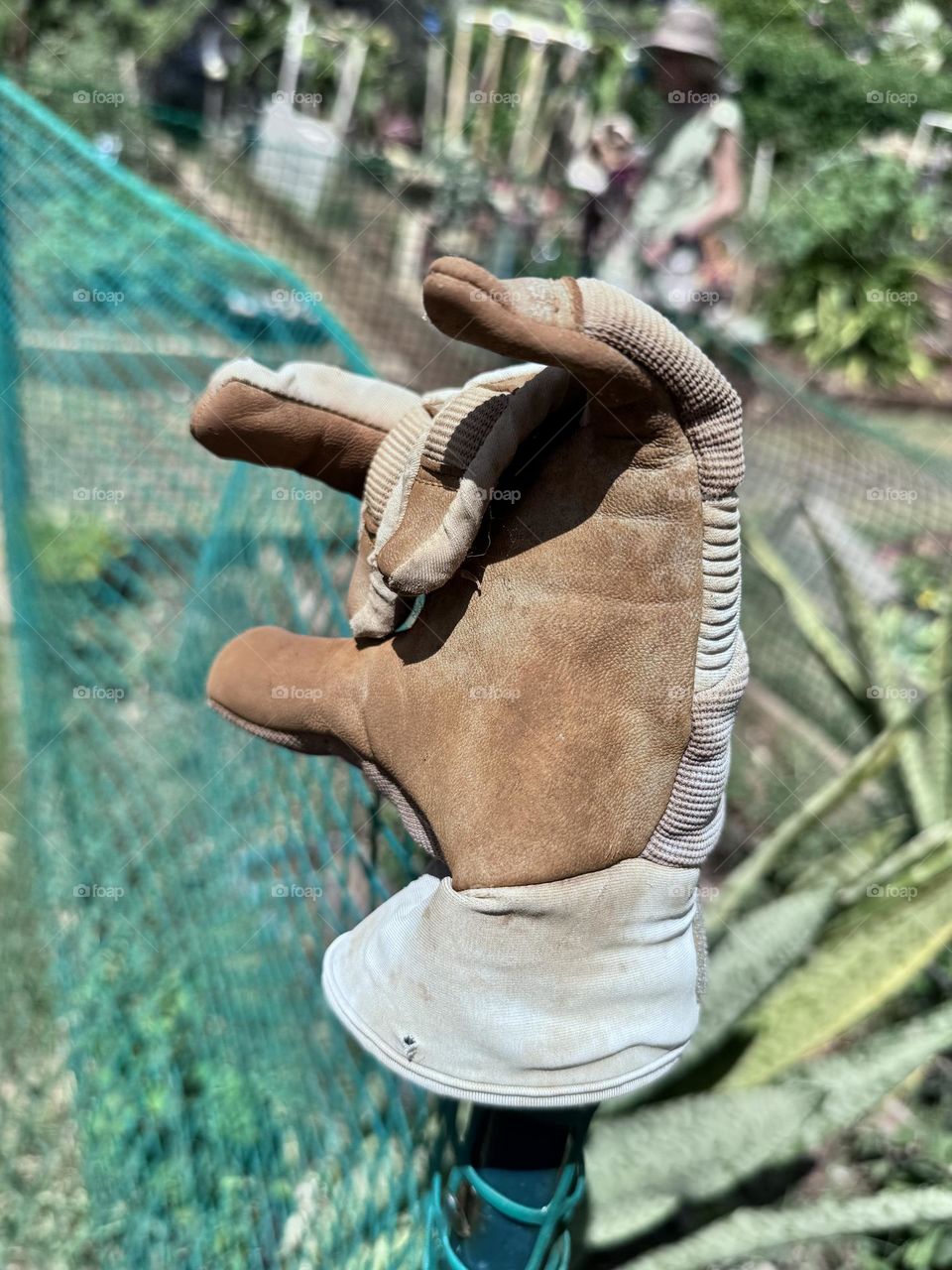 Friendly work glove on fence pole waves “hi” with sun hatted woman in the background of a verdant summer community garden 