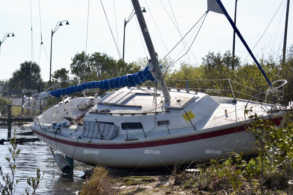 A white boat with a red stripe and a blue sail that’s shipwrecked on a riverbank 