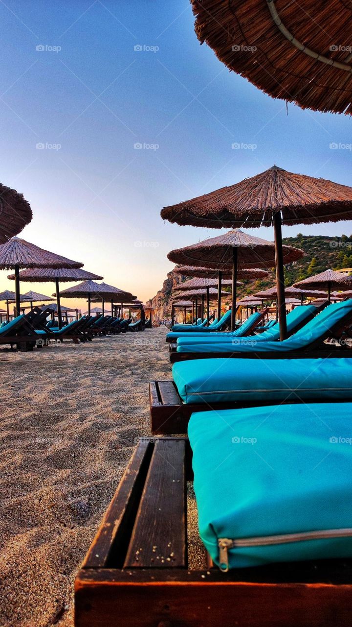 My absolute favourite place to travel to is the beach, with its beautiful views, sounds and calm presence, the sea is the perfect place for a vacation. A beautiful scenery of blue sunbeds, umbrellas and sandy beach during a sunset in Greece