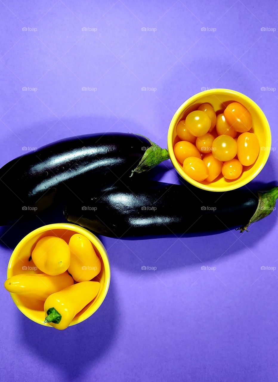 Yellow and purple complementary colours flatlay on a purple surface with eggplants, and yellow peppers and tomatoes in yellow bowls