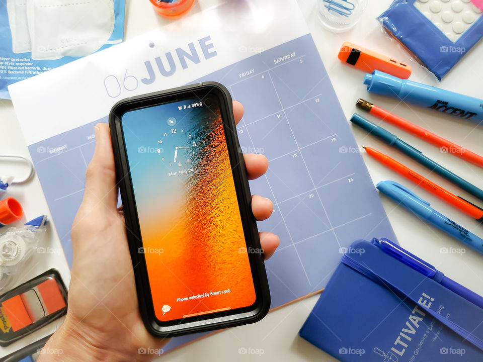 Blue and orange theme flatlay of a man holding his phone in front of a calendar surrounded by office supplies.