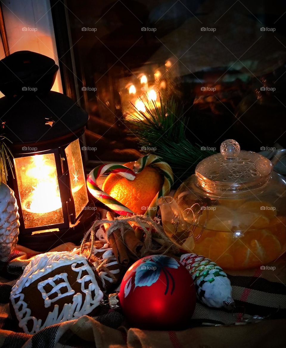 Waiting for Christmas. Evening. A burning candle in a lantern, gingerbread, tangerine, heart-shaped caramel canes, Christmas decorations and a teapot with tangerine tea. A candle is reflected in the window