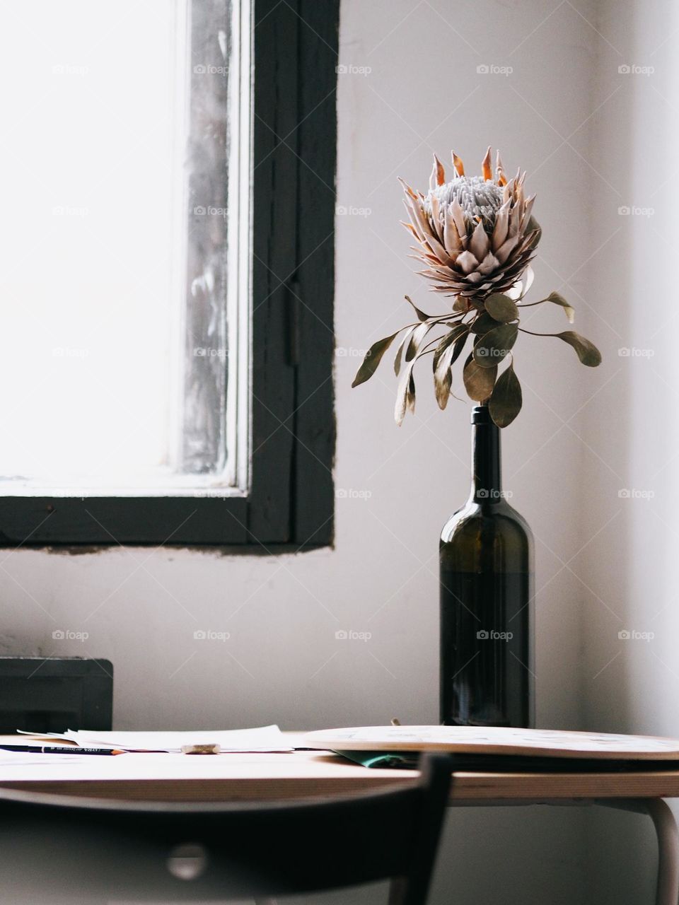 An isolated prothea flower stands in a glass bottle on a wooden table near a window, no people.