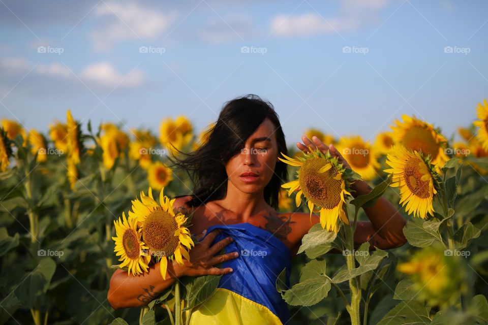 a beautiful, Ukrainian girl stands in a field of sunflowers, wrapped in a yellow-blue Ukrainian flag