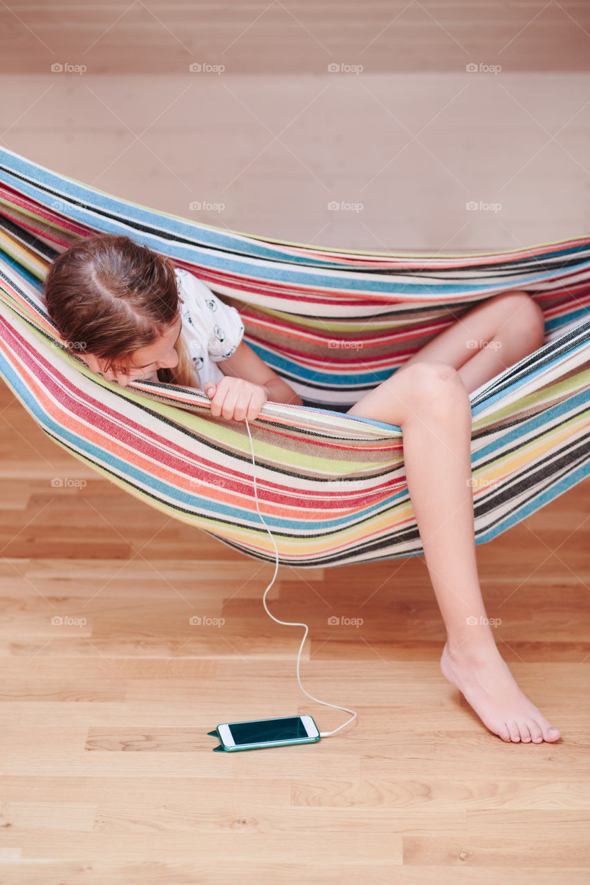 Teenage girl listening to music and chatting with friends on smartphone sitting in hammock at home
