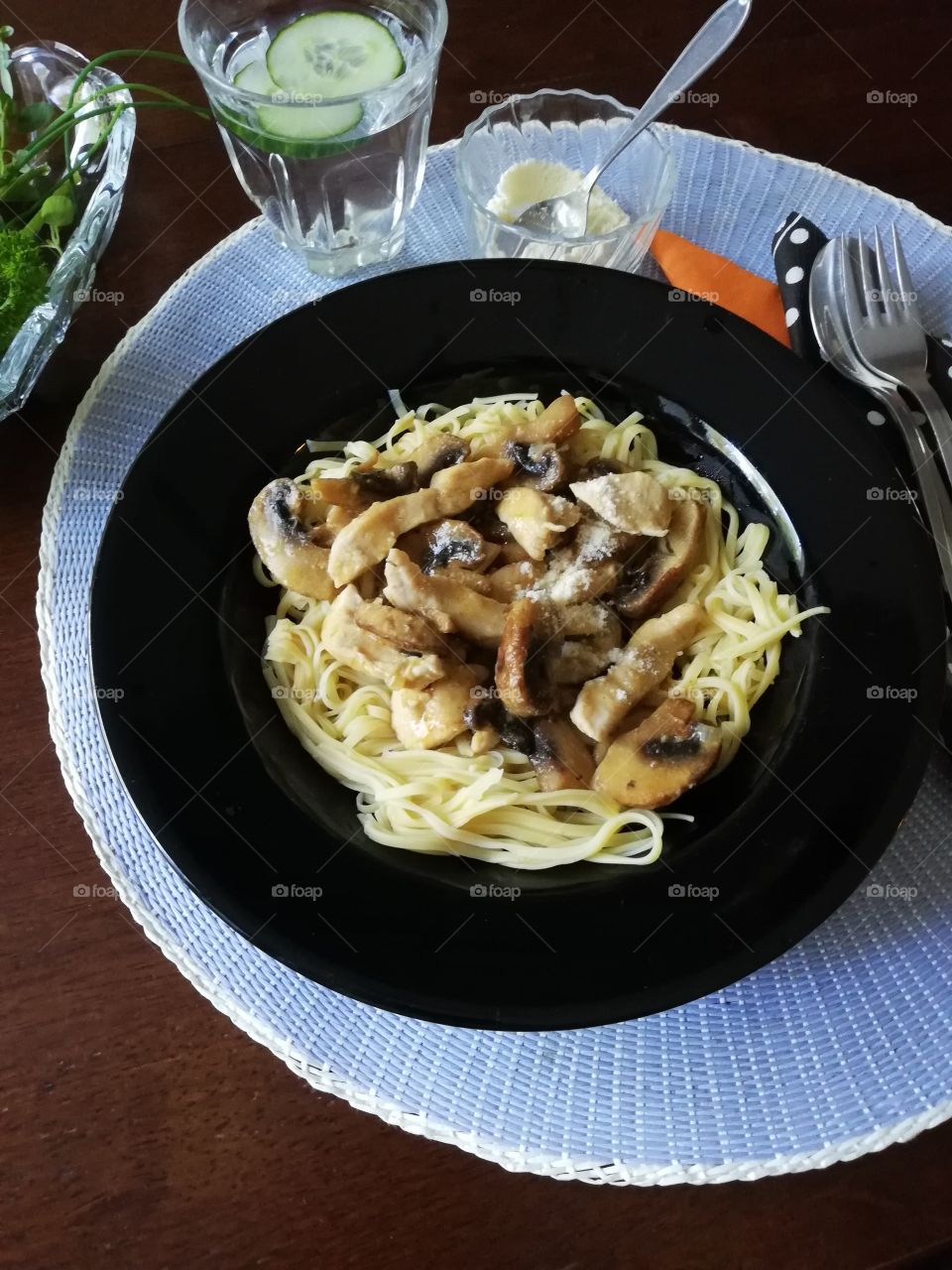 A black plate full of spagetti with chicken, mushrooms and parmesan on a light blue place mat. A glass of water with slices of cucumber and napkins, a fork, a spoon and a bowl of cheese on the table.