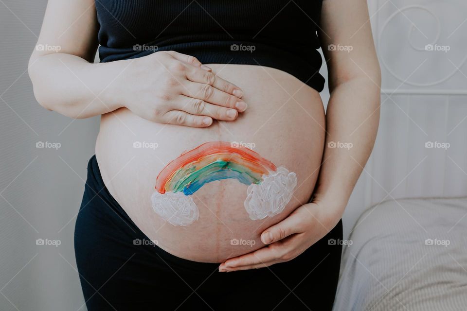 Portrait of one unrecognizable caucasian pregnant woman holding her belly with hands with painted acrylic paint rainbow with clouds,vtd side closeup. The concept of a new human.