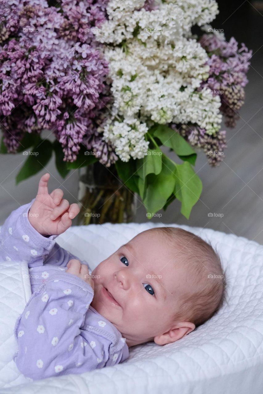 Little baby smiling in the cocoon and bouquet of white and violet lilacs