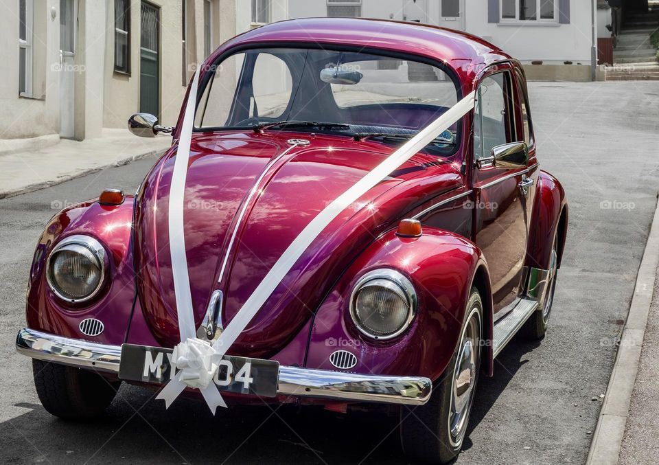 A pinky red VW Beetle wedding car, parked with white ribbon attached. 