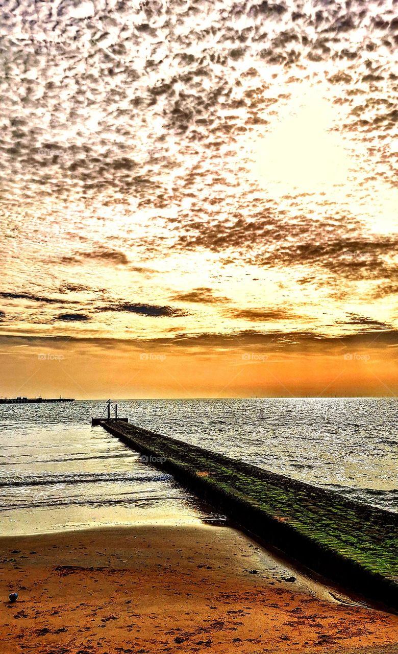 Sunrise over the sea with layers of mustard yellow and brown and a  stone jetty in the foreground extending to the horizon