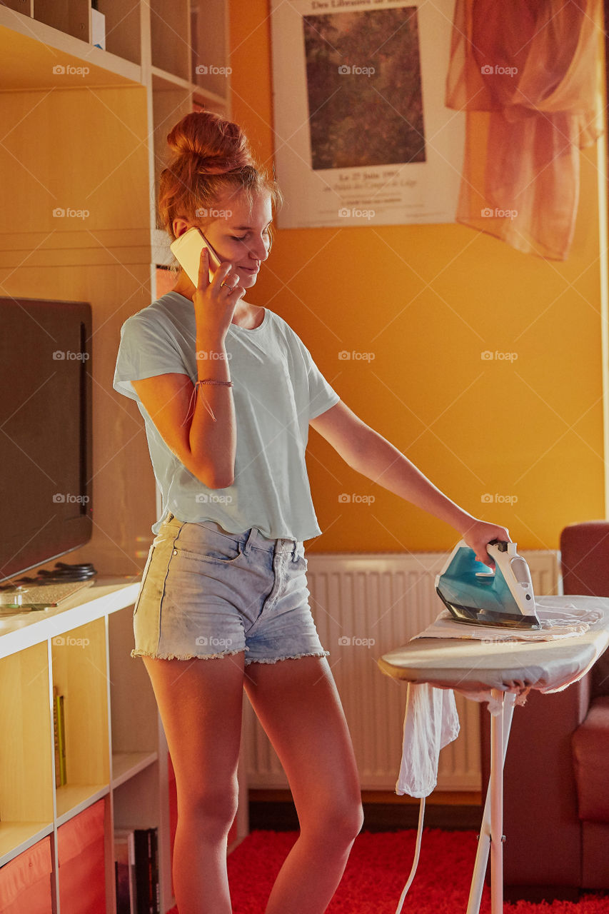 Young woman is ironing her clothes and talking on a smartphone simultaneously, standing by a ironing board in a room at home. Routine housekeeping task at home. Candid people, real moments, authentic situations