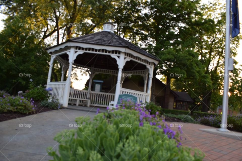Sidewalk leading up to gazebo in the late evening sunset, surrounded by trees and flowers. 