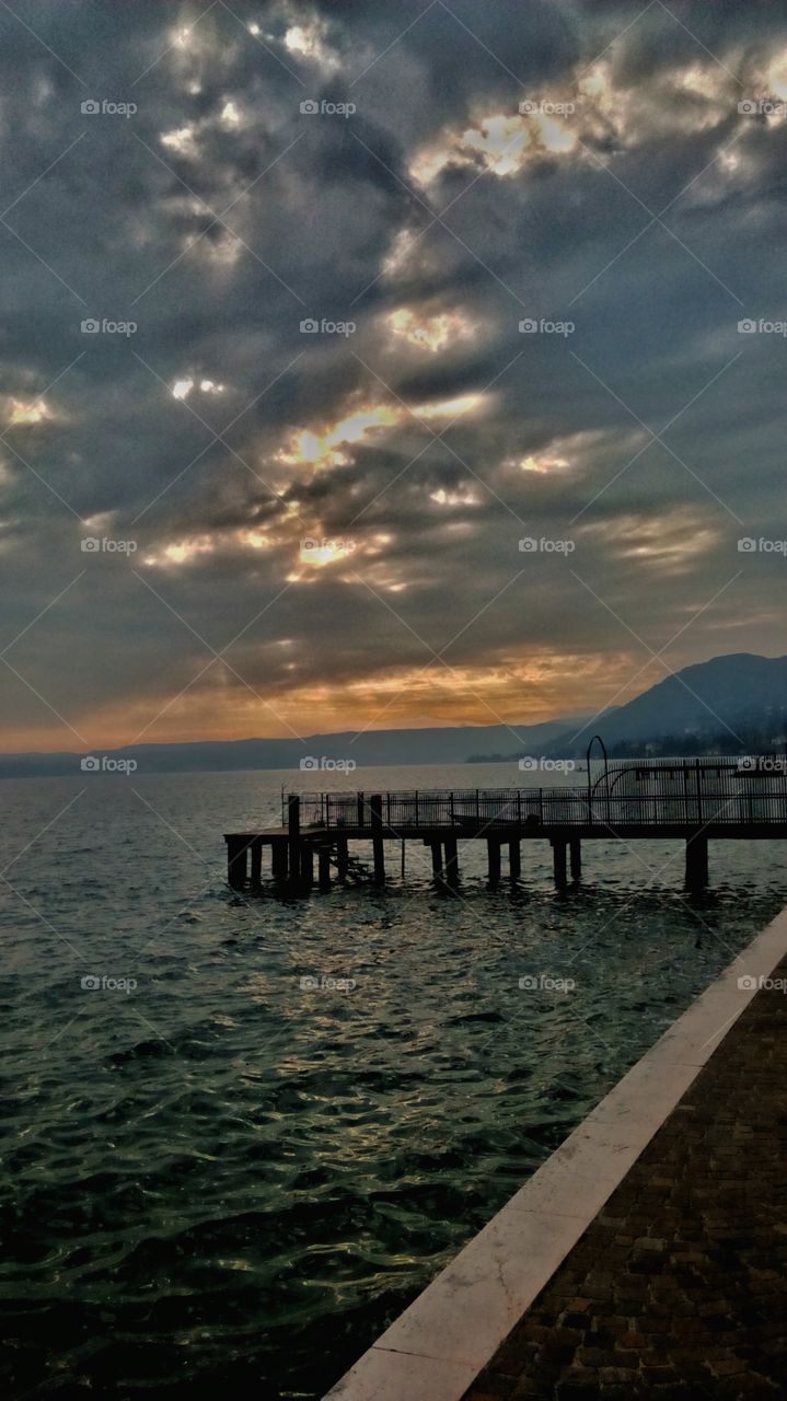 It is the sunrise over Lake Garda. In the background you can see the mountains surrounding the gulf, and the sky took colors and beautiful forms. The water with a bright blue, is driven by a light winter breeze. My personal view of a sunrise during a morning walk on the lakefront