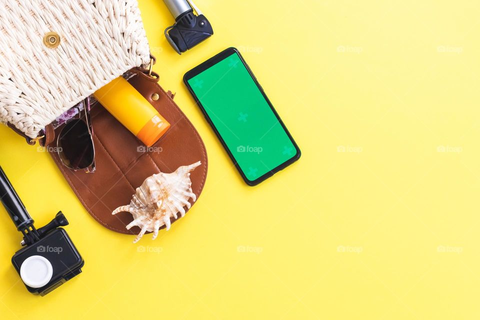 One straw bag with sunglasses spilling out of it, a small tube of sun cream, a shell, a camera and a mobile phone with a green screen lie on the left on a yellow background with copy space on the right, flat lay close-up.